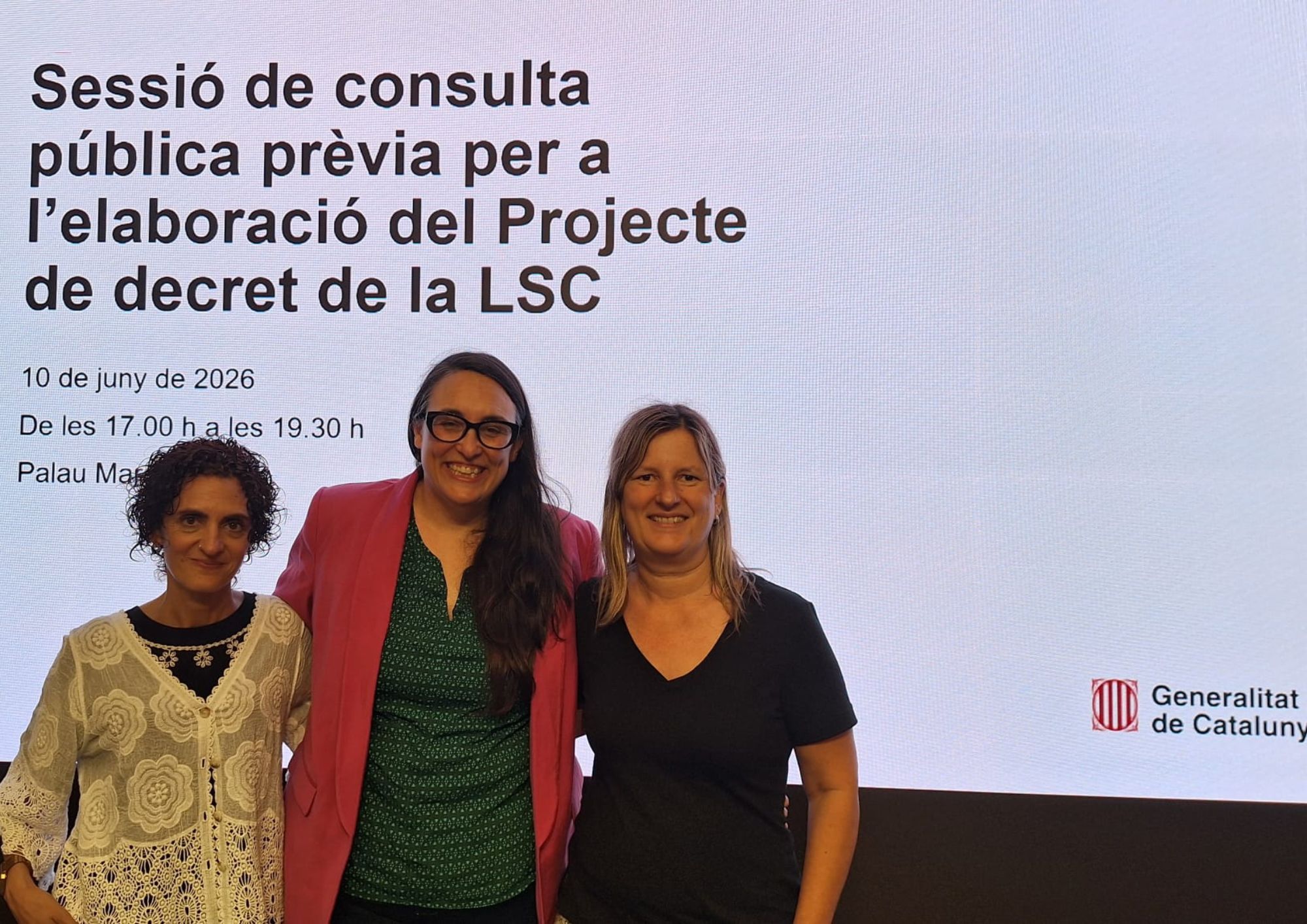 Les protagonistes: Esther Gálvez, Marta Bosch i Karen González somriuen en primer pla, posant juntes davant d'una gran pantalla. Mostren un ambient proper i de satisfacció per la jornada de treball. La pantalla de fons (Dades clau de l'acte): El text de la projecció detalla tota la informació institucional de la trobada: Títol: "Sessió de consulta pública prèvia per a l'elaboració del Projecte de decret de la LSC". Data i hora: 10 de juny de 2026, de les 17.00 h a les 19.30 h. Lloc: S'intueix l'inici del text "Palau Marc" (seu del Departament de Cultura / Política Lingüística). Logotip oficial: A la cantonada inferior dreta de la pantalla apareix clarament el logotip i el text de la Generalitat de Catalunya, confirmant el caràcter oficial de l'acte.
