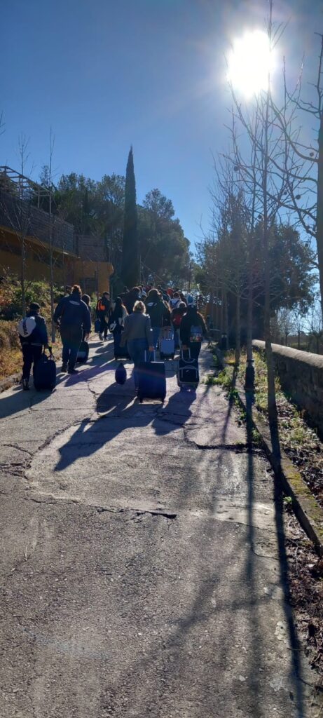 Un grup d'alumnes arrossegant les maletes per un camí asfaltat, fins a la casa de colònies. Fa bon dia.