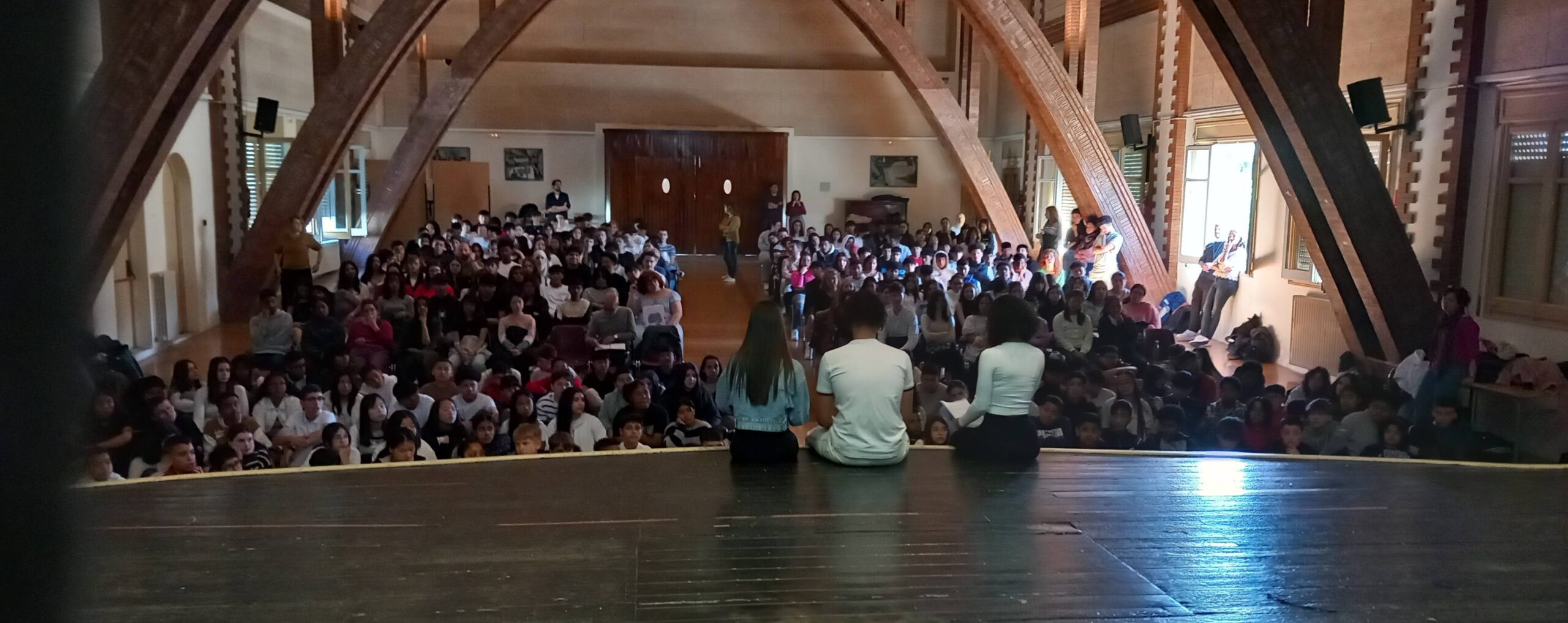 Sala d'actes de l'escola Cavall Bernat, plena d'alumnes. Els tres presentadors de l'acte, asseguts a la punta de l'escenari, adreçant-se al públic.