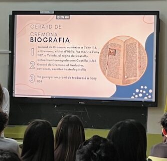 Es veu una pantalla digital il·luminada amb una presentació sobre Gerard de Cremona. Un nombrós grup d'alumnes observen la pantalla mentre 3 alumnes en presenten el contingut.