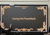 Es veu una pantalla digital il·luminada amb una presentació sobre Georg von Peuerbach. Un nombrós grup d'alumnes observen la pantalla mentre 3 alumnes en presenten el contingut.