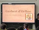 Es veu una pantalla digital il·luminada amb una presentació sobre Gerbert d'Orlhac. Un nombrós grup d'alumnes observen la pantalla mentre 3 alumnes en presenten el contingut.