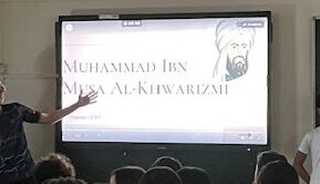Es veu una pantalla digital il·luminada amb una presentació sobre Muhammad ibn Musa al-Kwarizmi. Un nombrós grup d'alumnes observen la pantalla mentre 3 alumnes en presenten el contingut.