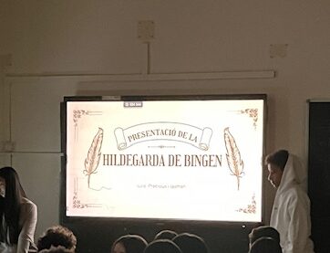 Es veu una pantalla digital il·luminada amb una presentació sobre Hildegarda de Bingen. Un nombrós grup d'alumnes observen la pantalla mentre 3 alumnes en presenten el contingut.