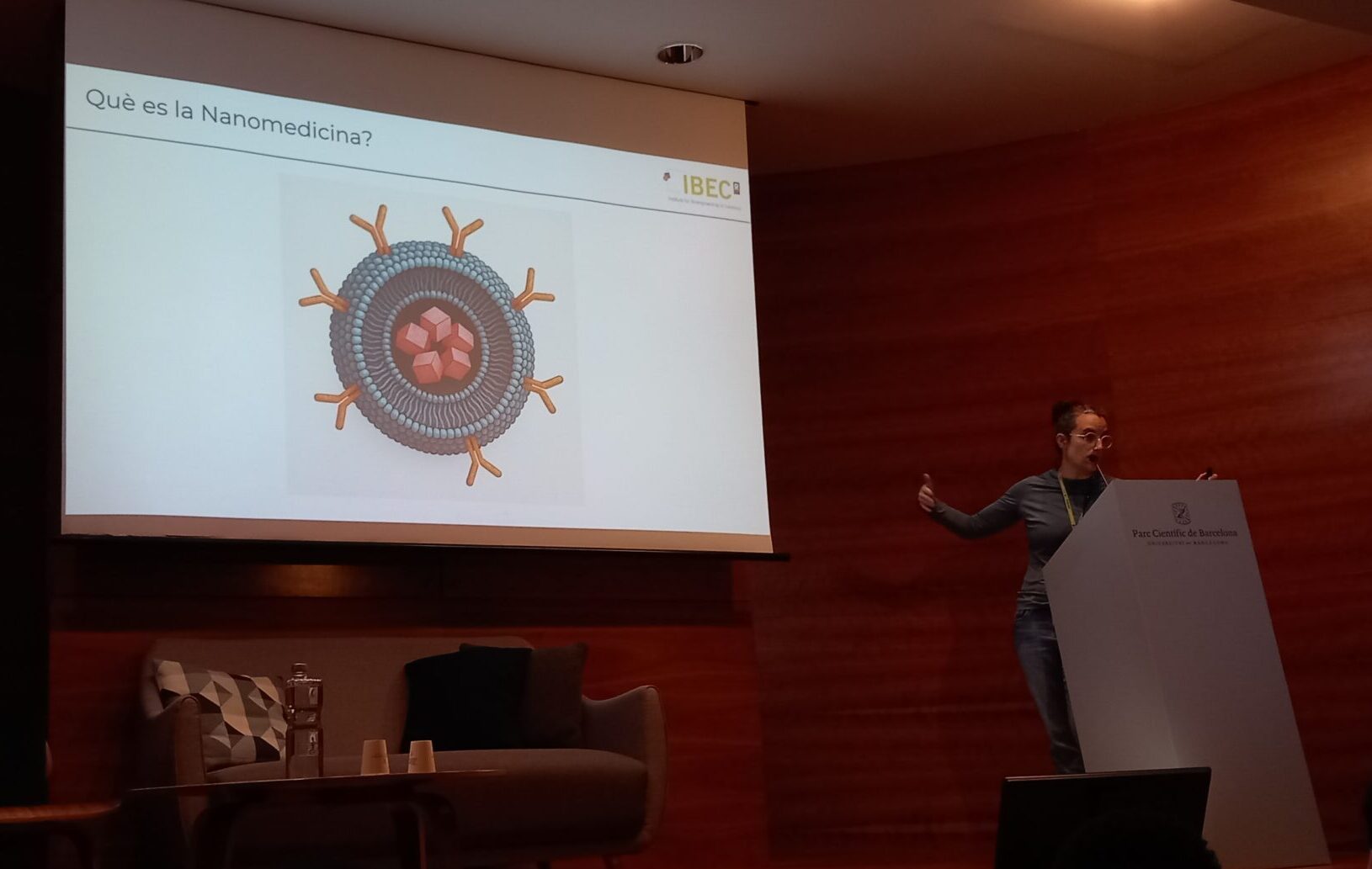 Una investigadora de l'IBEC parla des d'un faristol en un auditori, amb una diapositiva projectada a la pantalla que mostra una il·lustració d'una nanopartícula i la pregunta "Què es la Nanomedicina?".