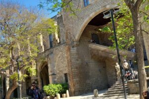 Vista exterior de l'antic Hospital de la Santa Creu de Barcelona en un dia assolellat: façana gòtica de pedra amb un gran arc de mig punt i una escala exterior, envoltada d'arbres amb fulles verdes. S'hi poden veure persones assegudes als bancs de la plaça i alumnes a les escales.