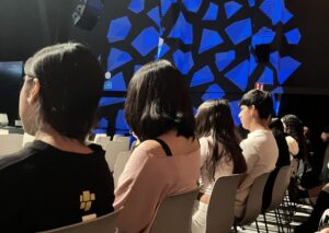 Alumnes de l'institut asseguts al pati de butaques del CCCB, esperant l'inici de la projecció. Al fons, una il·luminació escènica de formes geomètriques en blau decora l'espai de la sala.